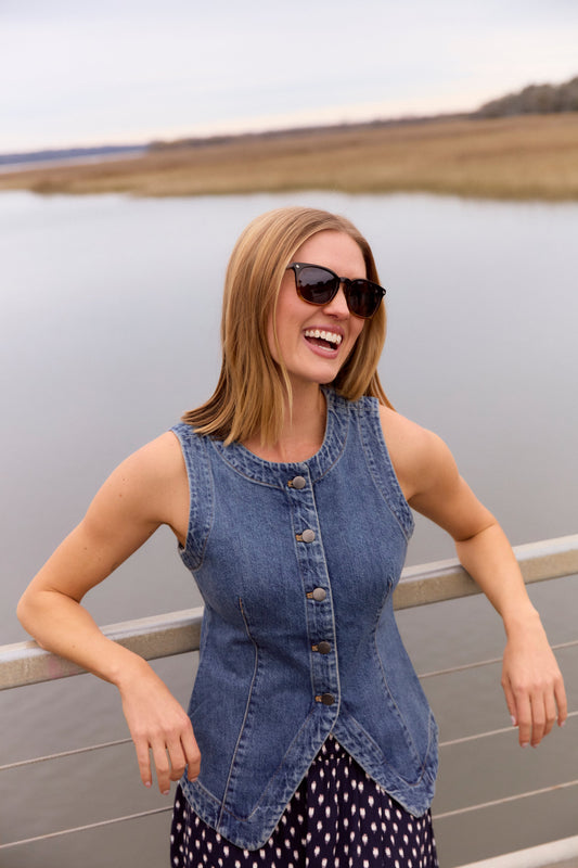woman in blue denim top in sunglasses against a railing by the lake