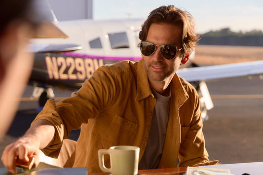 man in aviators at a table with a small plane on a runway in the background