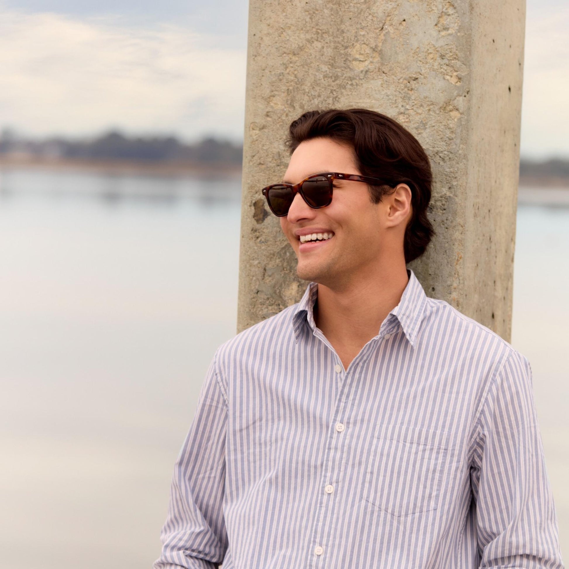 Lifestyle Image: Man wearing sunglasses and a striped shirt leaning against a concrete wall with water and sky in the background